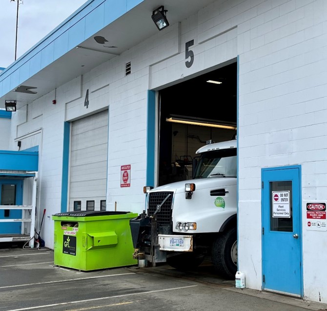 A white fleet building with blue trim has the front end of a garbage truck partially sticking out from its mechanic bay.