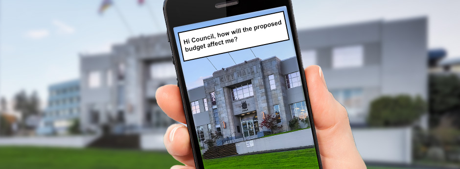 In front of a blurry image of Nanaimo's City Hall, a hand hold's a smart phone up with the question: Hi Council, how will the proposed budget affect me?