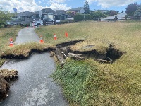 Grassy terrain adjacent to a walkway has caved in, revealing a circular concrete structure and pipes beneath the surface.