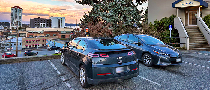 Two PHEV cars sit charging outside of Nanaimo City Hall, overlooking the orange sky and waterfront in the distance.