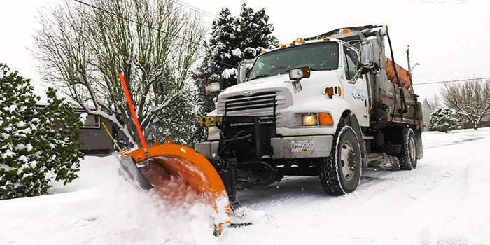A white truck with an orange plow pushes snow off of a snowy side road