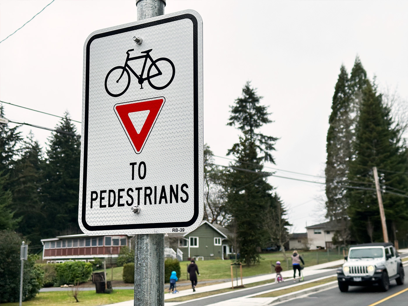 a cyclists yield to pedestrians sign with a parents and children walking and biking in the background