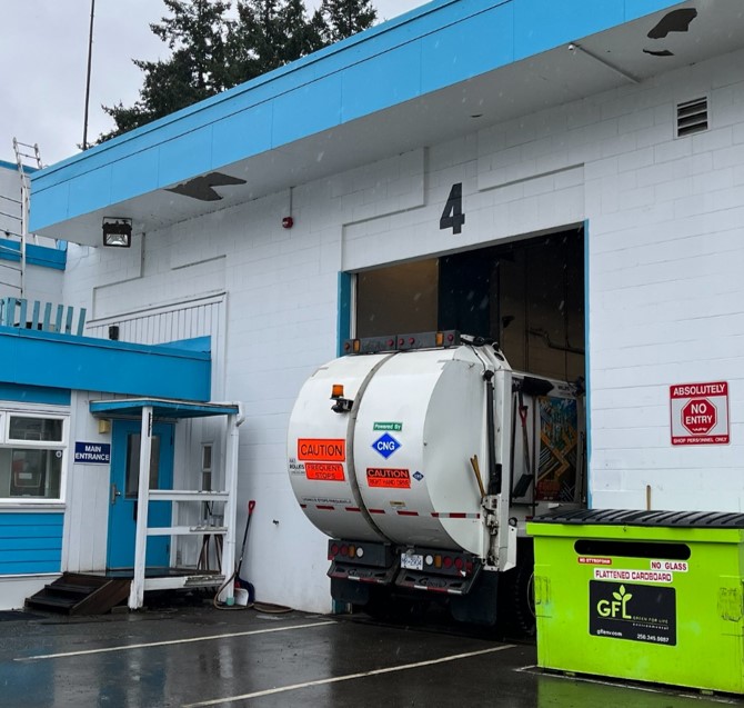 A white fleet building with blue trim has a garbage truck partially sticking out from its mechanic bay.