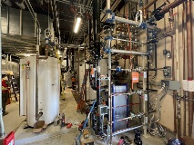 Alargestructureofpipesandvalvesfillstherightoftheframewhilealargehotwaterstoragetanktakesuptheleftsideoftheframe